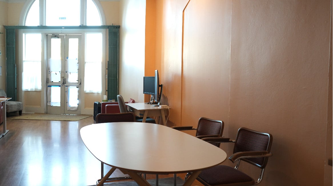 The main coworking room at Our Space Berkeley — round meeting table with Cesca chairs, arched front windows, hot desk with monitor